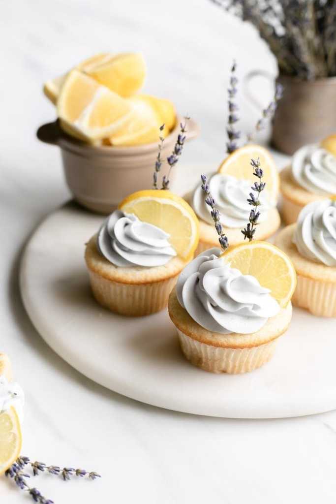 looking down at a platter of the lemon lavender cupcakes, with a bowl of lemon slices in the back as well as a vase with lavender