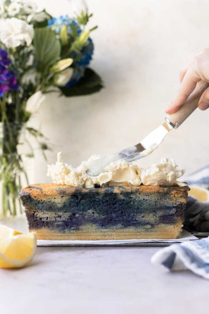 Spreading the white chocolate ganache onto the magic butterfly pea flower loaf cake, with a vase of blue, purple and white flowers in the background.