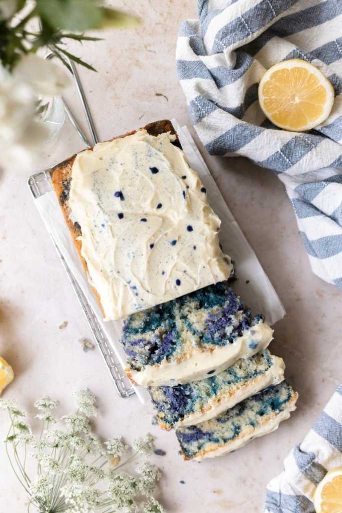 Flat lay of the butterfly pea flower loaf cake with three slices cut and laying on top of each other. Surrounding the cake are flowers, a blue and white striped napkin, and lemon slices.