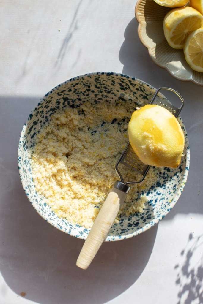 A zested lemon sitting on top of a zester, on the bowl with sugar and lemon zest.